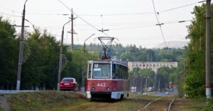 Трамвай Tatra T3 червоно-білого кольору з номером 4 на табло, бортовим номером 442, їде по рейках вздовж дороги, оточеної деревами. На задньому плані видно будівлю та червоний легковий автомобіль.