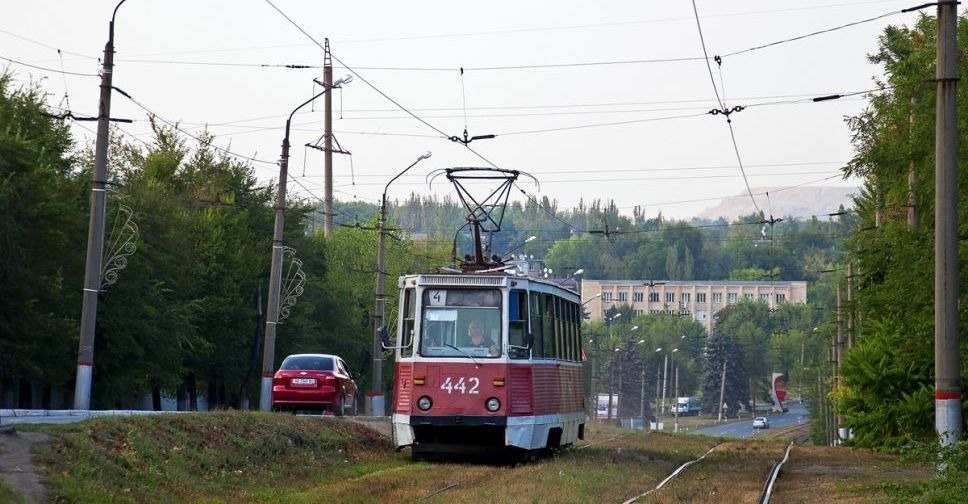 Трамвай Tatra T3 червоно-білого кольору з номером 4 на табло, бортовим номером 442, їде по рейках вздовж дороги, оточеної деревами. На задньому плані видно будівлю та червоний легковий автомобіль.